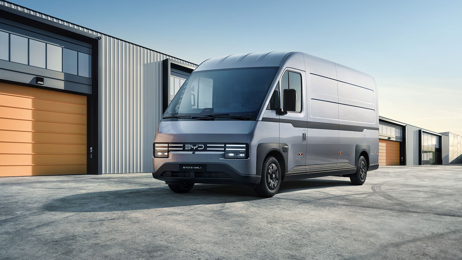 Ασημί ηλεκτρικό van BYD E-Vali μπροστά από σύγχρονες βιομηχανικές εγκαταστάσεις.